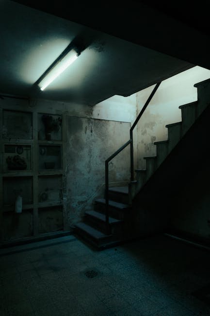 Interior view of a basement flat with a dimly lit staircase featuring wooden steps and a metal handrail, ascending from a concrete floor to an upper level. The surrounding walls are unfinished, with visible patches of peeling paint and exposed plaster. To the left, there is a white shelving unit with multiple compartments holding various items, possibly cleaning supplies or household objects. The ceiling has a fluorescent light fixture providing minimal illumination, casting shadows across the space. The environment suggests a conversion or storage area within a property, typical of a basement flat during a home relocation process. This image is relevant to professional removals and furniture transport services, such as those offered by Man with Van Charing Cross, involved in packing, moving, and logistical planning for house moves, including navigating staircases and tight corners.