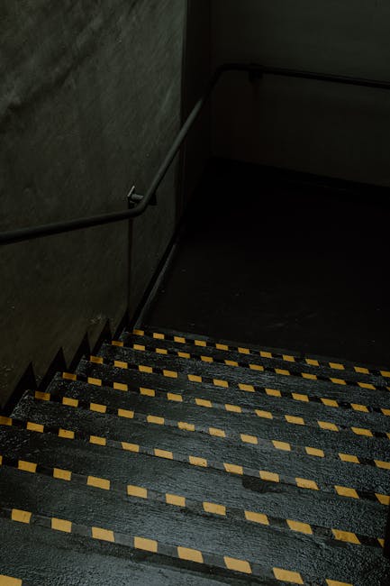 A dimly lit staircase inside a residential building, featuring black-painted steps with yellow and beige strip markings along the edges for safety and visibility. A metal handrail runs along the left side, attached to dark textured wall surfaces. The staircase appears narrow, with several steps leading downward, indicating a basement or cellar level. The environment suggests an area often used for moving household items, as part of a home relocation process, with no furniture or boxes visible on these stairs. The lighting is sparse, emphasizing the utilitarian nature of the staircase. This setting reflects the internal logistics involved in house removals, supporting services such as furniture transport and packing during relocation, which are handled professionally by companies like Man with Van Charing Cross.