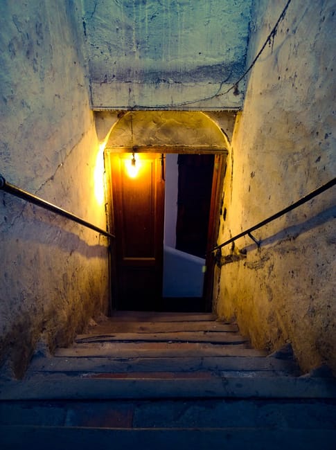 Interior view of a basement flat with a dimly lit staircase featuring wooden steps and a metal handrail, ascending from a concrete floor to an upper level. The surrounding walls are unfinished, with visible patches of peeling paint and exposed plaster. To the left, there is a white shelving unit with multiple compartments holding various items, possibly cleaning supplies or household objects. The ceiling has a fluorescent light fixture providing minimal illumination, casting shadows across the space. The environment suggests a conversion or storage area within a property, typical of a basement flat during a home relocation process. This image is relevant to professional removals and furniture transport services, such as those offered by Man with Van Charing Cross, involved in packing, moving, and logistical planning for house moves, including navigating staircases and tight corners.