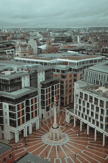 An aerial view of a densely built urban area featuring modern multi-storey office buildings surrounding a large, circular paved square with a prominent white monument in the centre. The monument consists of a tall, slim column topped with a decorative element, and there are a few pedestrians walking across the square and along the pavements between the buildings. In the background, the cityscape extends with additional office blocks, historic buildings, and a cloudy sky overhead. This scene captures the central part of Charing Cross vicinity, highlighting the architecture and layout typical of a city undergoing home relocation and furniture transport activities, as seen from above during daylight hours.