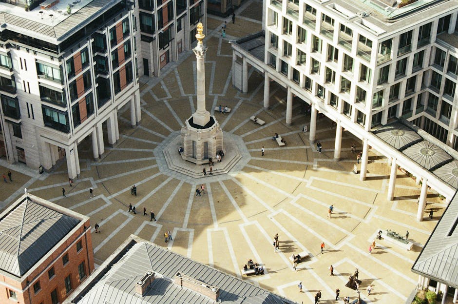 An aerial view of Golden Square in Charing Cross, showcasing a centrally located monument with a tall column topped by a statue, surrounded by a paved circular area with geometric red and beige brick patterns. Modern office buildings and residential flats with large windows and balconies encircle the square. In the lower part of the image, a frontline building with a grey metal roof and brick facade is visible. The area appears well-maintained with pedestrians walking along the pathways. This urban scene provides context for house removals and urban furniture transport, highlighting the importance of careful planning during a home relocation. Man with Van Charing Cross may operate in such city environments, ensuring efficient packing, loading, and transport of household items through narrow streets or open public spaces, supporting logistics for moving services requested at [PAGE_TITLE].