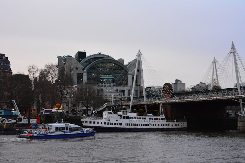 A photograph taken near The Savoy hotel in Charing Cross shows a river scene with two boats docked along the bank. The larger boat is a white passenger vessel with multiple windows, appearing to be in the process of being loaded or unloaded as part of a house removal or relocation service by Man with Van Charing Cross. The smaller boat, painted blue and white, is positioned closer to the foreground and is stationary. Behind the boats, there is a bridge with tall, white suspension cables and pylons, spanning across the river. The background features the modern, glass-fronted structure of the Royal Festival Hall, with a partly overcast sky overhead, casting diffuse lighting over the scene. The environment suggests a relocating operation involving furniture and equipment being transported via river, consistent with professional removals and packing services offered by Man with Van Charing Cross near the Savoy.