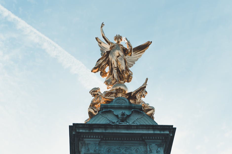 The image depicts a tall, ornate monument with a gilded top featuring a detailed statue of an angel or winged figure, positioned above a decorative pedestal. The statue appears to be made of metal with a gold finish, contrasting against the clear, blue sky in the background. The structure below the statue is constructed from stone or concrete, with intricate carvings and architectural details. The scene is captured during daylight, with natural lighting highlighting the metallic shine of the statue and the textures of the monument. This setting relates to the context of central London, near Charing Cross, and reflects the historical and cultural significance of the area. In the context of house removals and transport services, such monuments indicate a well-visited urban environment where professional removals, such as those provided by Man with Van Charing Cross, often operate, ensuring safe and efficient logistics for relocating belongings amidst busy city landmarks.