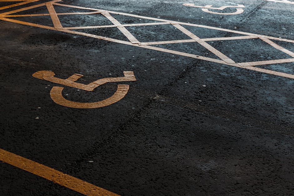 Close-up of a designated accessible parking space marked with a yellow wheelchair symbol painted on the dark asphalt surface outside a building. Adjacent to the parking space, there are white painted 