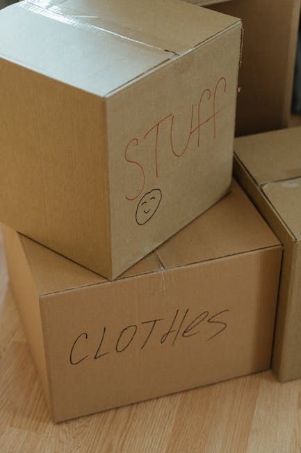 Close-up view of two brown cardboard moving boxes stacked inside a home, with the top box labeled 'STUFF' in red marker and a simple smiley face drawn below, and the lower box labeled 'CLOTHES' also in red marker. The boxes are sealed with packing tape, placed on a wooden floor, and surrounded by additional boxes, suggesting an ongoing packing or home relocation process. The setting indicates interior space prepared for furniture transport and house removals, consistent with professional moving services provided by Man with Van Charing Cross. Proper packing materials such as cardboard and tape are visible, emphasizing careful packing and transit preparation for a moving project.