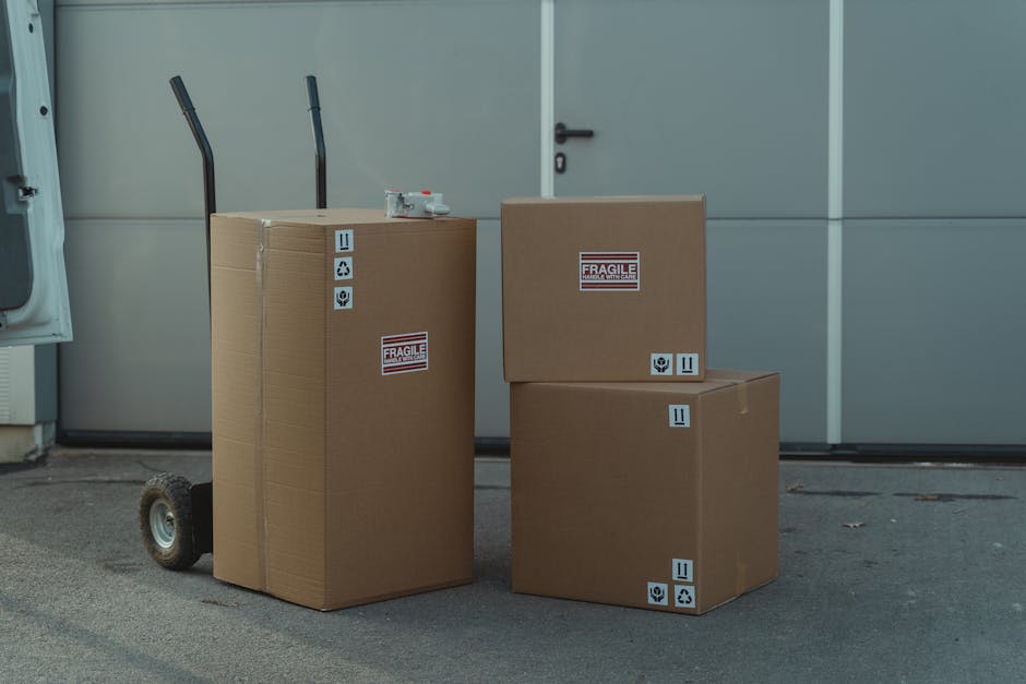Four cardboard moving boxes of varying sizes, sealed with packing tape, are placed on an outdoor pavement in front of a large gray garage door. Two boxes are positioned on a manual hand trolley with black handles and small wheels, while the remaining two are directly on the ground. The boxes feature printed labels indicating fragility, handling instructions, and recycling symbols. The scene suggests the process of home relocation, with the boxes ready for transport as part of furniture transport and packing and moving activities managed by Man with Van Charing Cross. Natural daylight illuminates the area, highlighting the organized stacking and careful packing preparations typical during the loading process in house removals.