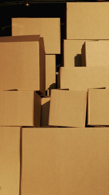 Close-up view of two brown cardboard moving boxes stacked inside a home, with the top box labeled 'STUFF' in red marker and a simple smiley face drawn below, and the lower box labeled 'CLOTHES' also in red marker. The boxes are sealed with packing tape, placed on a wooden floor, and surrounded by additional boxes, suggesting an ongoing packing or home relocation process. The setting indicates interior space prepared for furniture transport and house removals, consistent with professional moving services provided by Man with Van Charing Cross. Proper packing materials such as cardboard and tape are visible, emphasizing careful packing and transit preparation for a moving project.