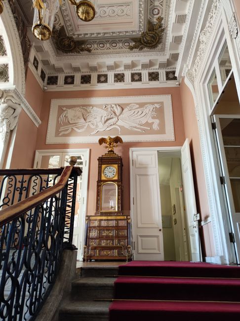 The interior view of a grand, historic building shows a section of a staircase with a decorative wrought iron handrail and a red carpet runner, leading up to a landing. Above the staircase, the ceiling features ornate molding and a decorative painting. An antique wooden clock with a gold eagle finial is situated on a small, ornate marble-topped chest at the top of the stairs, against a wall with a large, classical relief sculpture of reclining figures painted in soft beige tones. Large windows with white frames allow natural light to fill the space, highlighting the elegant architectural details. This setting is part of a high-ceilinged entrance hall or foyer, typical of traditional house removals or home relocation projects, as conducted by Man with Van Charing Cross, which involves careful handling and protection of the building’s historic interior features during furniture transport and packing processes.