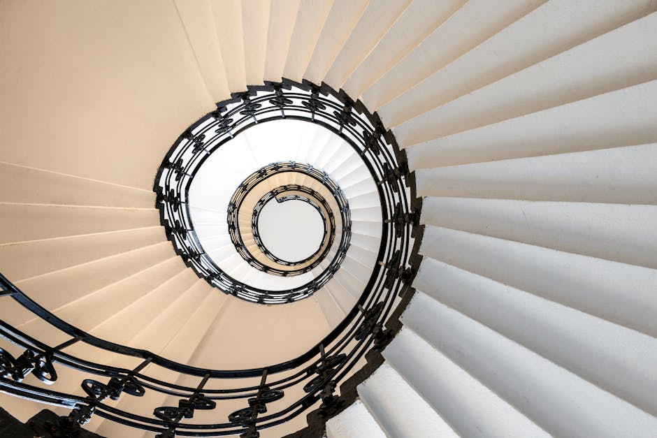 A view looking down a winding staircase with beige steps and a black wrought iron railing that spirals downward in a circular pattern. The staircase is located inside a building and is well-lit, showing the smooth surface of the steps and the decorative design of the railing. The image captures the interior environment where the stairs are part of a classic or historic property, relevant to house relocation and moving services. This perspective is often seen during the loading process of furniture or packing materials, which might be in the vicinity outside or nearby, although no objects or moving equipment are visible in this particular shot. Man with Van Charing Cross occasionally handles furniture transport and packing in properties with Georgian staircases, focusing on careful protection of furniture during home relocations.