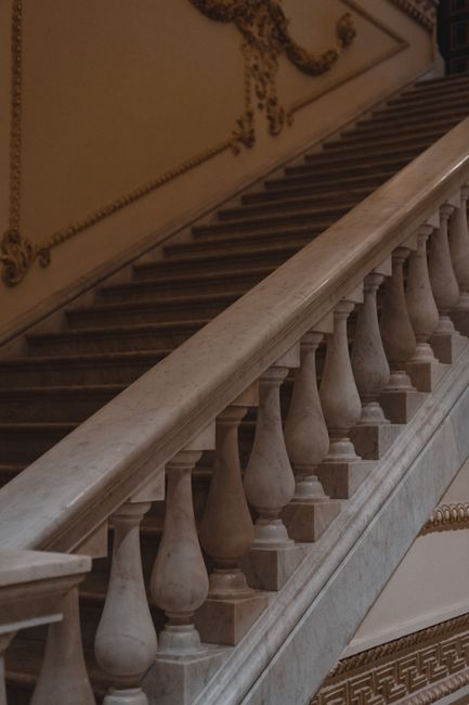 The interior view of a grand, historic building shows a section of a staircase with a decorative wrought iron handrail and a red carpet runner, leading up to a landing. Above the staircase, the ceiling features ornate molding and a decorative painting. An antique wooden clock with a gold eagle finial is situated on a small, ornate marble-topped chest at the top of the stairs, against a wall with a large, classical relief sculpture of reclining figures painted in soft beige tones. Large windows with white frames allow natural light to fill the space, highlighting the elegant architectural details. This setting is part of a high-ceilinged entrance hall or foyer, typical of traditional house removals or home relocation projects, as conducted by Man with Van Charing Cross, which involves careful handling and protection of the building’s historic interior features during furniture transport and packing processes.