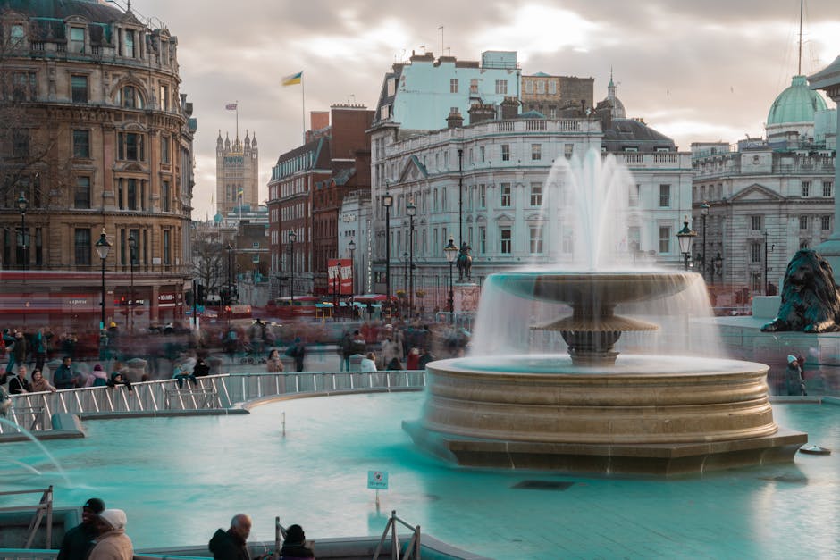 The image shows Trafalgar Square in London during daytime, with a large decorative fountain prominently in the foreground. The fountain features a wide, round stone basin with cascading water above, and several water jets are visible, creating a misty effect. Surrounding the fountain, there are numerous people walking and sitting on benches, some wearing coats and hats, suggesting cool weather. In the background, historic and modern buildings, including a large white classical-style building with columns and decorative sculptures, as well as other structures with varying architectural styles, line the square. A flag is flying on a flagpole atop one of the buildings. The scene is illuminated by soft, natural light, possibly during late afternoon, with a partly cloudy sky overhead. This setting illustrates a typical busy day at one of London's iconic landmarks where local residents and tourists are engaged in sightseeing, walking, or relaxing in the area, often involved in home relocation or furniture transport activities as part of moving services by companies such as Man with Van Charing Cross.