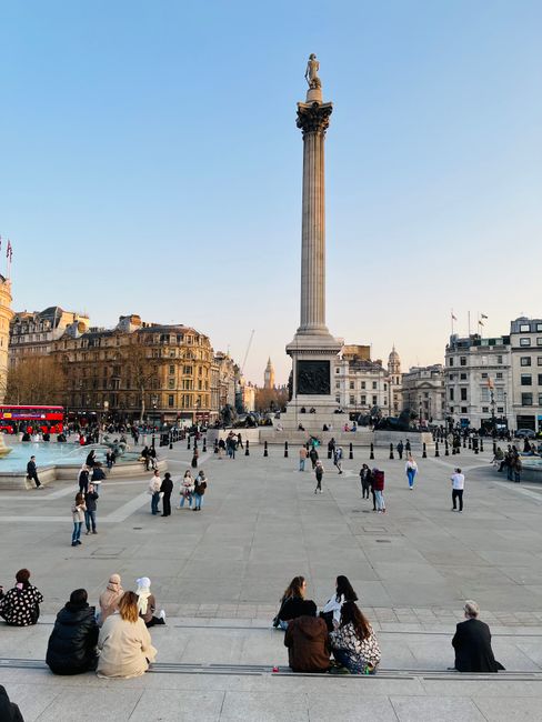 A wide view of Trafalgar Square showing a tall stone column with a statue on top, surrounded by historic buildings with intricate facades. In the foreground, several groups of people are seated on steps and walking across the paved open space. Some individuals are carrying cardboard boxes and wrapped furniture, suggesting ongoing home relocation or furniture transport. A loading area near the base of the column shows a man with a trolley and moving blankets, engaged in loading boxes or belongings onto a van. The setting is outdoors during daylight with clear blue skies, and the scene captures the process of packing and moving within a busy public space, with the backdrop of iconic London architecture. The image reflects the logistical aspects of removals and the importance of coordinated transport for house moves, provided by companies like Man with Van Charing Cross.