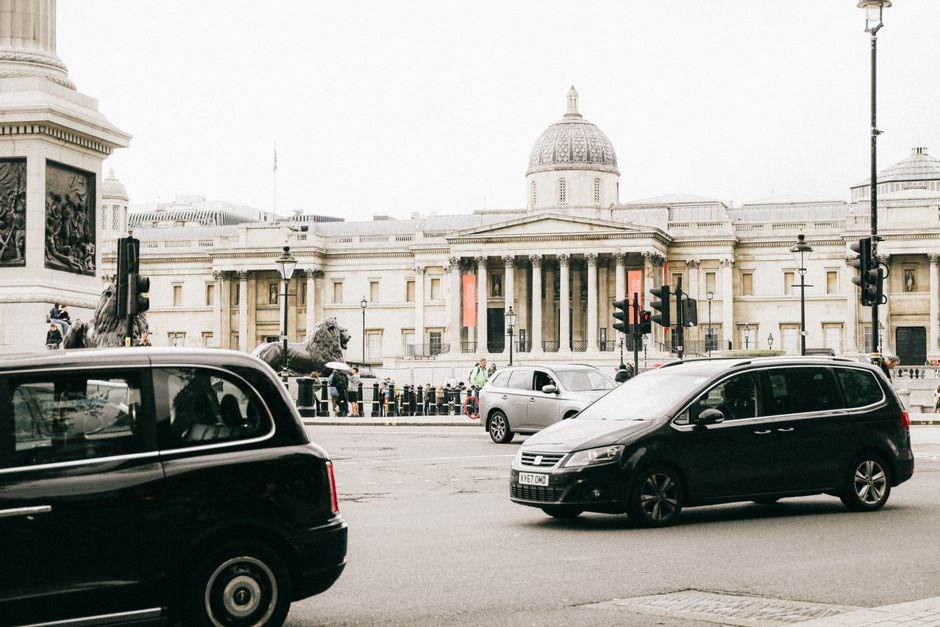 A busy street scene in front of a historic building with classical architectural details, such as columns and a domed roof, situated in Trafalgar Square. In the foreground, a black van and a black MPV are seen parked or moving along the road, with traffic lights and street lamps visible. Behind the vehicles, people are lined up near the building, some standing and others walking, with a few police officers present. The area appears to be a designated loading bay or street parking zone, with barrier posts and signage indicating parking rules. The environment is well-lit with natural daylight, and the overall scene captures the typical urban logistics involved in house removals and furniture transport services, as provided by Man with Van Charing Cross, supporting efficient packing and moving operations near historic landmarks.