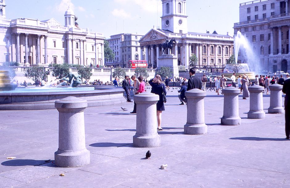 A wide view of Trafalgar Square showing a tall stone column with a statue on top, surrounded by historic buildings with intricate facades. In the foreground, several groups of people are seated on steps and walking across the paved open space. Some individuals are carrying cardboard boxes and wrapped furniture, suggesting ongoing home relocation or furniture transport. A loading area near the base of the column shows a man with a trolley and moving blankets, engaged in loading boxes or belongings onto a van. The setting is outdoors during daylight with clear blue skies, and the scene captures the process of packing and moving within a busy public space, with the backdrop of iconic London architecture. The image reflects the logistical aspects of removals and the importance of coordinated transport for house moves, provided by companies like Man with Van Charing Cross.
