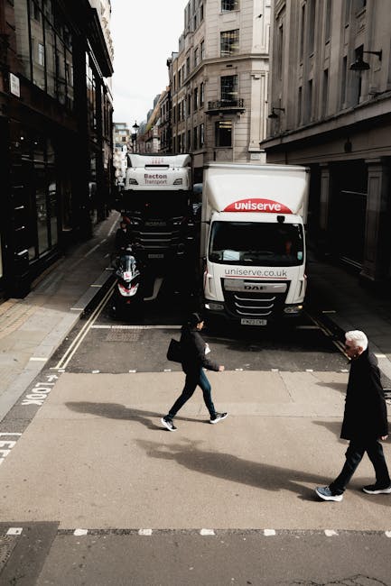 A street scene on a narrow urban road with tall buildings on either side showing a house removal process. Two temporary parked trucks belonging to Uniserve, a removals company, are positioned on the street, with one truck partially blocking the road and a motorcycle parked in front of them. The trucks are white with red and black branding, and they appear to be loaded with packing materials or furniture for an imminent home relocation. In the foreground, a woman in dark clothing carrying a bag walks across a pedestrian crosswalk, while a man in a suit and white hair follows behind her. The environment is well-lit with natural daylight, highlighting the busy logistical activity related to furniture transport and packing during a moving service, consistent with the services described on the webpage about urgent same-day removals in WC2 facilitated by Man with Van Charing Cross.
